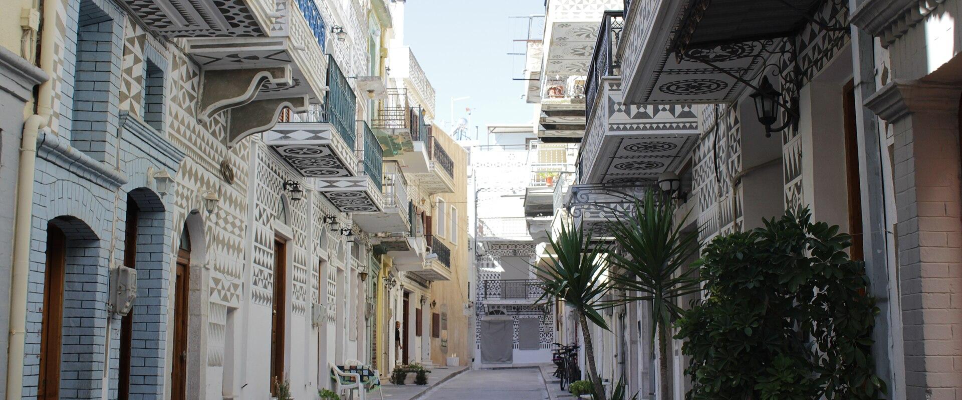 A narrow street in a village featuring buildings with distinctive black and white geometric patterns (xysta) on the facades and balconies.