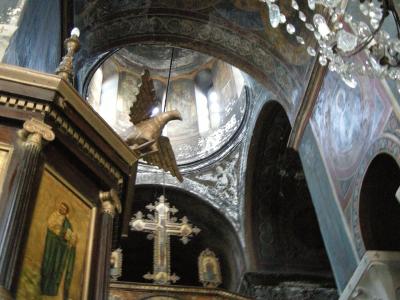 The interior of an Orthodox church featuring a wooden iconostasis with religious icons, a hanging brass eagle lectern, a large cross, and frescoed walls and ceilings under a crystal chandelier.