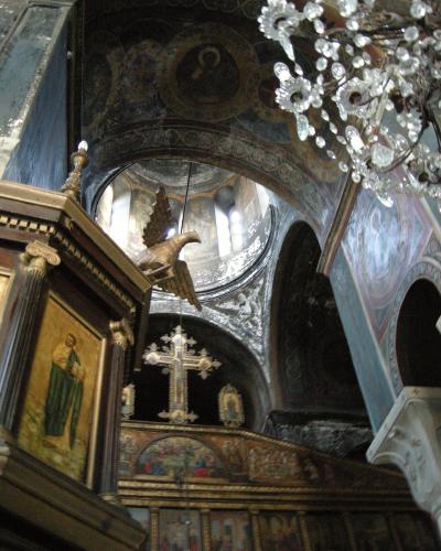 The interior of an Orthodox church featuring a wooden iconostasis with religious icons, a hanging brass eagle lectern, a large cross, and frescoed walls and ceilings under a crystal chandelier.
