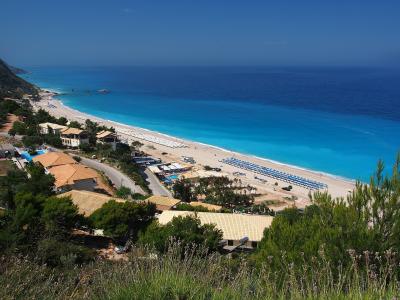 A high-angle view of a long, sandy beach with turquoise water, lined with rows of blue umbrellas and sunbeds, with buildings and a road in the foreground.