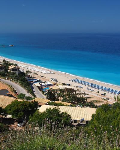 A high-angle view of a long, sandy beach with turquoise water, lined with rows of blue umbrellas and sunbeds, with buildings and a road in the foreground.