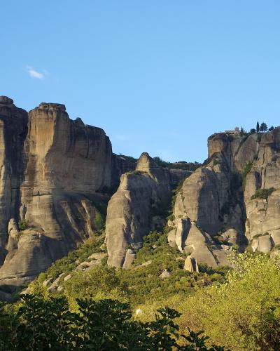 A landscape photograph showing towering sandstone rock pillars with a monastery complex perched on the summit of the formation on the right side, set against a blue sky.