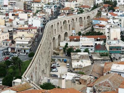 A high-angle photograph showing a massive, two-tiered stone aqueduct with numerous arches running diagonally through a densely built urban area with terracotta-roofed buildings.