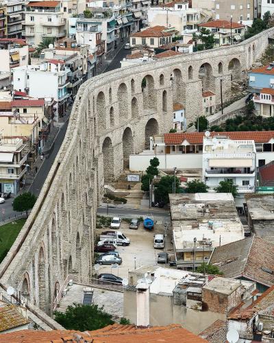 A high-angle photograph showing a massive, two-tiered stone aqueduct with numerous arches running diagonally through a densely built urban area with terracotta-roofed buildings.
