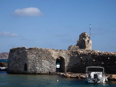 A stone Venetian castle ruin situated directly on the water's edge with a Greek flag flying from a tower and a small white boat moored in the foreground.