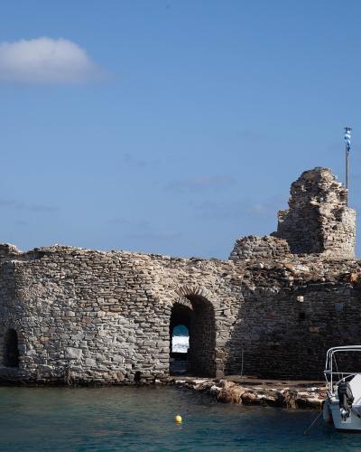 A stone Venetian castle ruin situated directly on the water's edge with a Greek flag flying from a tower and a small white boat moored in the foreground.