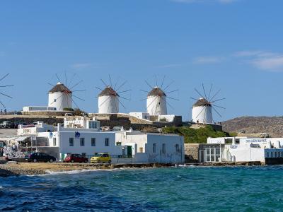 A row of five iconic white windmills with conical thatched roofs and large wooden sails, situated on a hill overlooking the sea and a coastal road with parked cars and tourists.