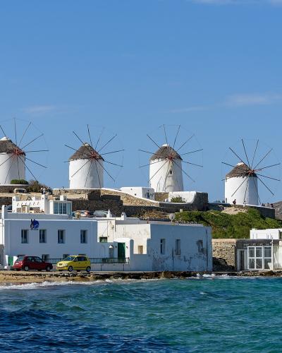A row of five iconic white windmills with conical thatched roofs and large wooden sails, situated on a hill overlooking the sea and a coastal road with parked cars and tourists.