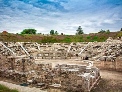 The image shows the stone ruins of an ancient Greek theatre with tiered seating, a circular orchestra area, and a red-domed structure visible on the hill in the background.