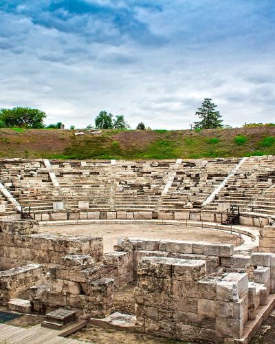 The image shows the stone ruins of an ancient Greek theatre with tiered seating, a circular orchestra area, and a red-domed structure visible on the hill in the background.