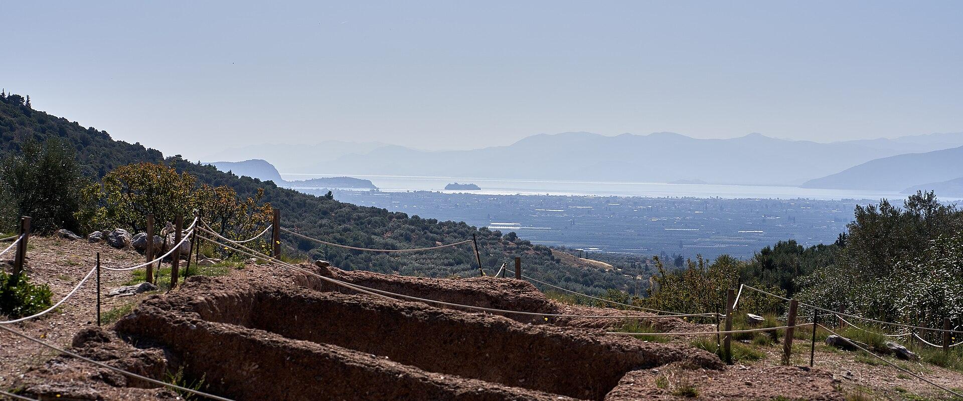 A view from an elevated archaeological site showing excavated earthworks or trenches in the foreground, overlooking a coastal plain and a large body of water with distant mountains.