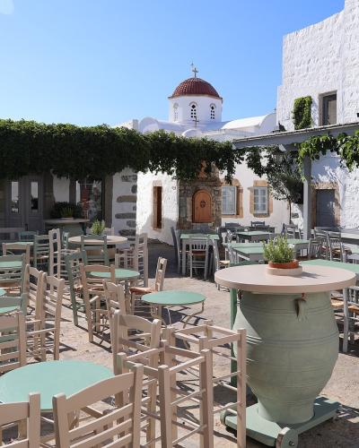 An outdoor cafe terrace with light green tables and wooden chairs, shaded by a vine-covered pergola, with a white-washed building and a church dome visible in the background.