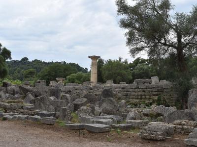The image shows the ruins of an ancient Greek temple site with scattered stone blocks and a single standing column, set against a backdrop of green trees and hills.
