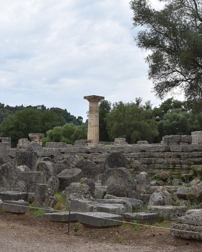 The image shows the ruins of an ancient Greek temple site with scattered stone blocks and a single standing column, set against a backdrop of green trees and hills.