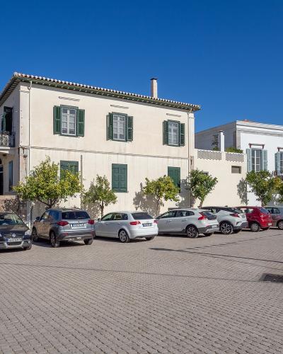 A paved public square or parking area in a Greek town, featuring a large two-story cream building with green shutters, a row of parked cars, and a black lamppost with Greek flags.