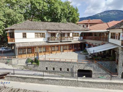 The image shows a traditional stone and wood building with a slate roof, featuring a ground-floor shop with wooden shutters and an upper-level balcony, situated in a paved square with a stone retaining wall in the foreground.