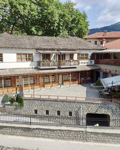 The image shows a traditional stone and wood building with a slate roof, featuring a ground-floor shop with wooden shutters and an upper-level balcony, situated in a paved square with a stone retaining wall in the foreground.