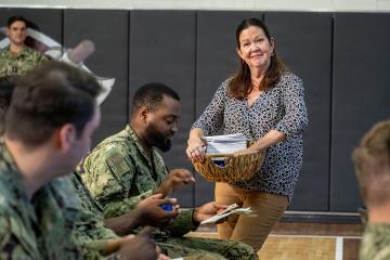 A woman in civilian clothing stands holding a basket of papers while interacting with several US Navy sailors in camouflage uniforms inside a gymnasium with padded walls.