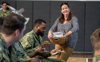 A woman in civilian clothing stands holding a basket of papers while interacting with several US Navy sailors in camouflage uniforms inside a gymnasium with padded walls.