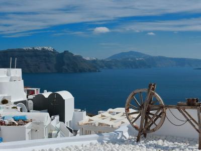 A view of white-washed buildings on a cliffside overlooking the deep blue Aegean Sea and the volcanic caldera islands in the distance, with a large wooden wheel structure in the foreground.
