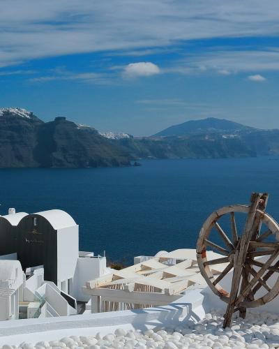 A view of white-washed buildings on a cliffside overlooking the deep blue Aegean Sea and the volcanic caldera islands in the distance, with a large wooden wheel structure in the foreground.