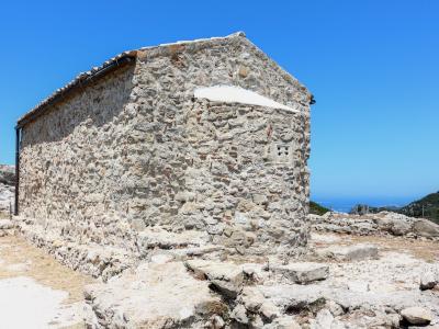 A small, rectangular stone building with a tiled roof and whitewashed accents, situated on a rocky, elevated terrain with a view of the sea and distant hills in the background.