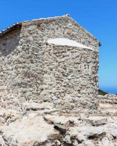 A small, rectangular stone building with a tiled roof and whitewashed accents, situated on a rocky, elevated terrain with a view of the sea and distant hills in the background.