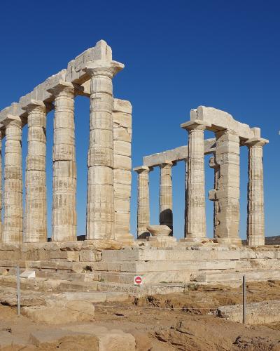 The image shows the ancient ruins of a Doric temple with standing columns and a partial entablature, situated on a rocky coastal promontory under a clear blue sky.