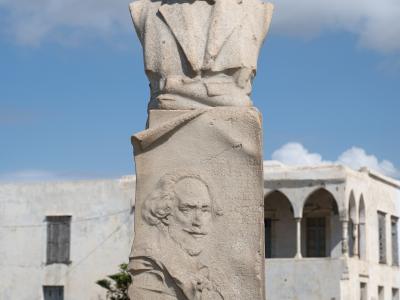 A stone bust monument of a man with a mustache standing outdoors with white buildings in the background.