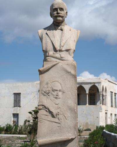 A stone bust monument of a man with a mustache standing outdoors with white buildings in the background.