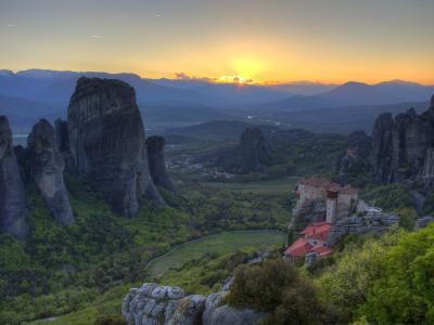 A landscape photograph showing towering sandstone rock pillars rising above a green valley, with a monastery built on a cliff edge in the foreground and a sunset in the background.