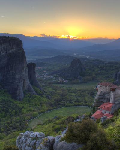 A landscape photograph showing towering sandstone rock pillars rising above a green valley, with a monastery built on a cliff edge in the foreground and a sunset in the background.