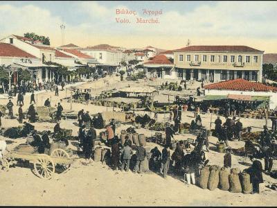 A vintage colorized postcard showing a bustling outdoor market square with vendors, crowds, horse-drawn carts, and surrounding buildings with red-tiled roofs.