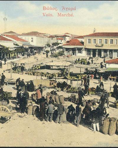 A vintage colorized postcard showing a bustling outdoor market square with vendors, crowds, horse-drawn carts, and surrounding buildings with red-tiled roofs.