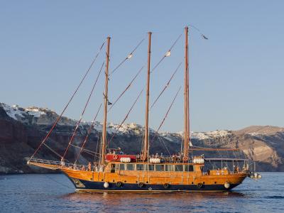 A large wooden three-masted sailing vessel with passengers on deck, sailing on calm blue water with the white-washed cliffside buildings of a Greek island caldera in the background.