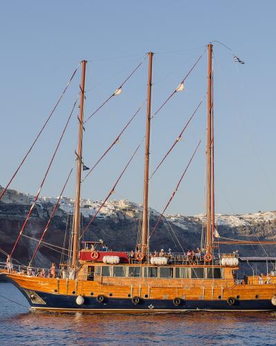 A large wooden three-masted sailing vessel with passengers on deck, sailing on calm blue water with the white-washed cliffside buildings of a Greek island caldera in the background.