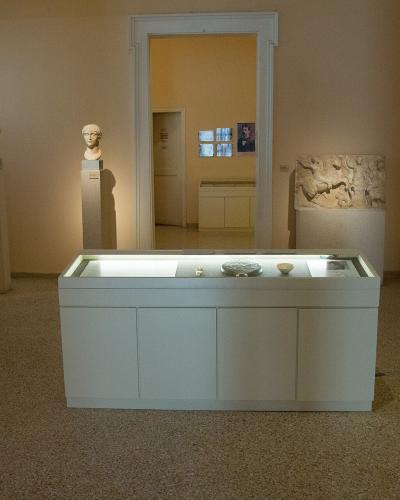 An interior view of a museum gallery featuring classical sculptures, including busts on pedestals and a relief, displayed around a central glass-topped case.