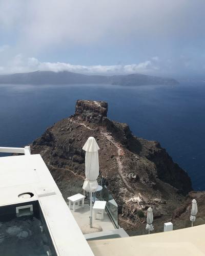 A view from a white terrace with a pool and closed umbrellas looking out over the caldera towards a prominent, jagged volcanic rock formation jutting into the sea.
