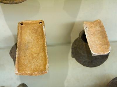 A close-up photograph of two ancient marble artifacts, identified as Cycladic "palettes," displayed inside a glass museum case on supports.