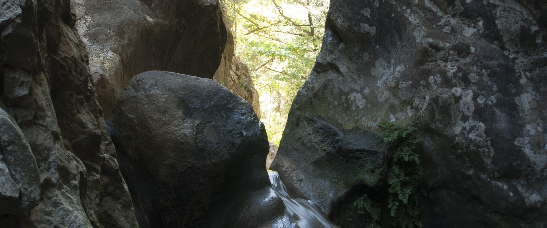 A small waterfall cascading down a smooth rock face into a pool, framed by large boulders and trees in a narrow gorge.