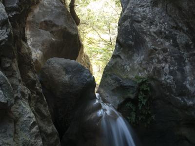 A small waterfall cascading down a smooth rock face into a pool, framed by large boulders and trees in a narrow gorge.
