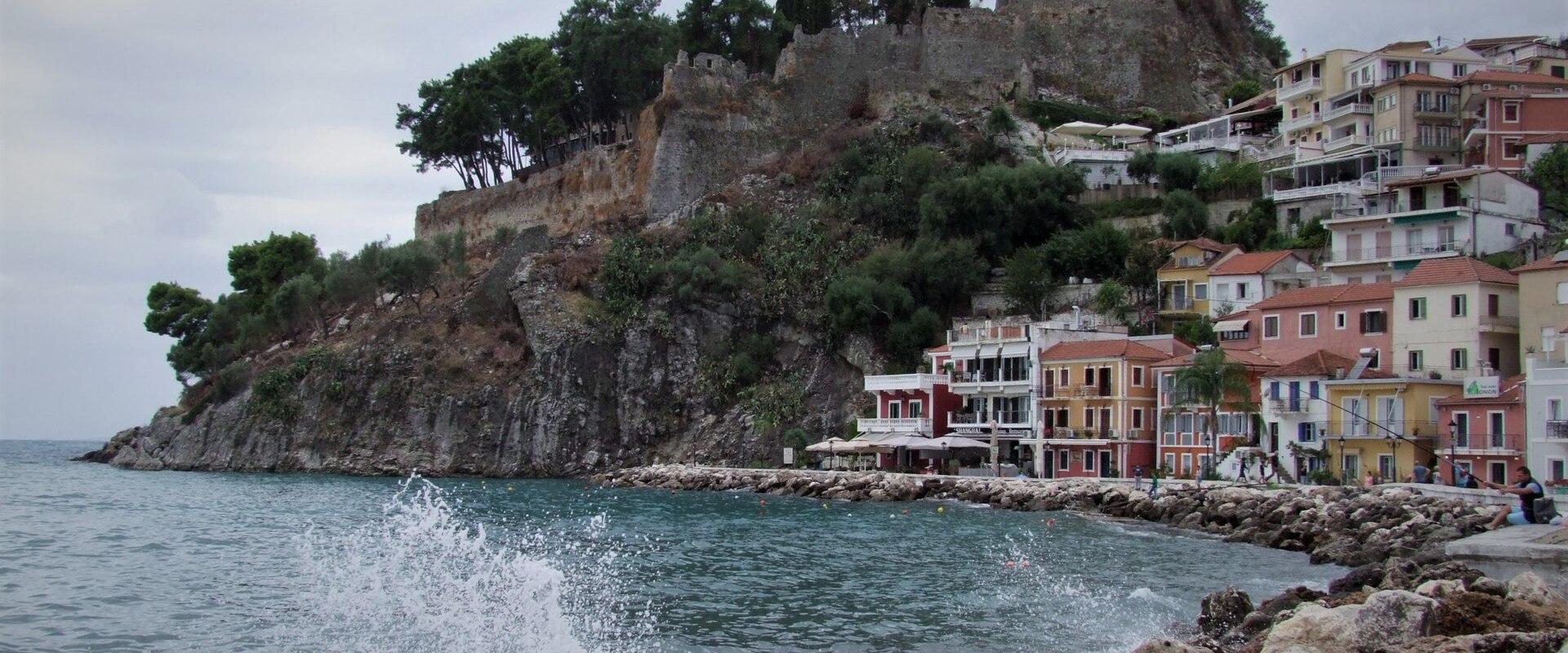 The image shows a coastal scene with a rocky promenade in the foreground, colorful buildings along the shore, and a fortified structure on a hilltop overlooking the sea.