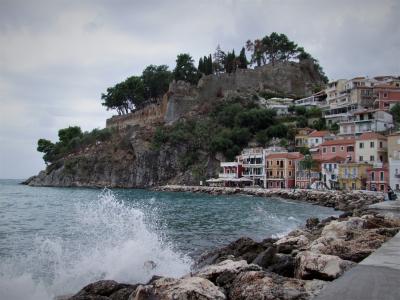 The image shows a coastal scene with a rocky promenade in the foreground, colorful buildings along the shore, and a fortified structure on a hilltop overlooking the sea.