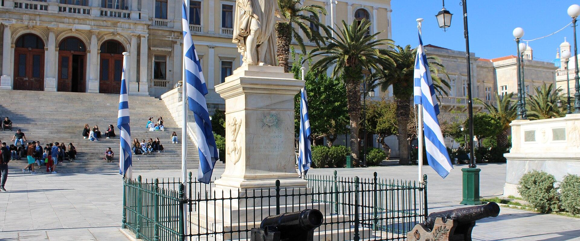 A large Neoclassical building with a grand staircase, a central statue on a pedestal surrounded by Greek flags and cannons, and people sitting on the steps.