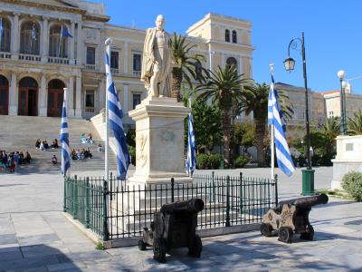 A large Neoclassical building with a grand staircase, a central statue on a pedestal surrounded by Greek flags and cannons, and people sitting on the steps.