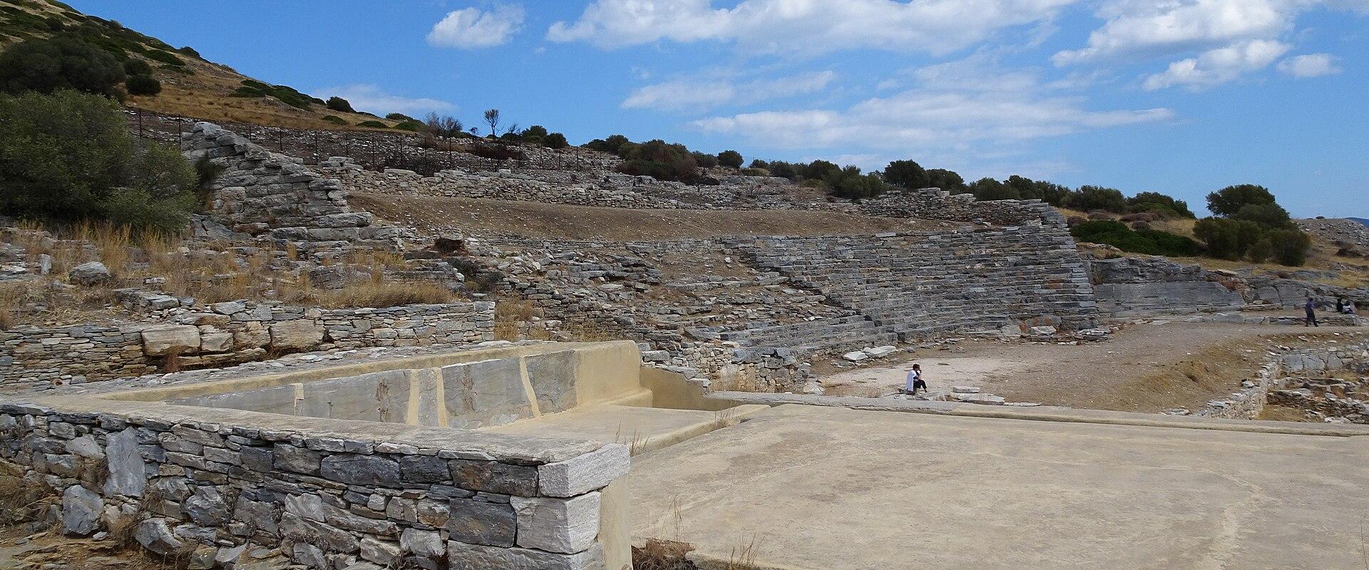 The image shows the ruins of an ancient stone theater with tiered seating carved into a hillside and a reconstructed concrete stage area in the foreground under a blue sky.