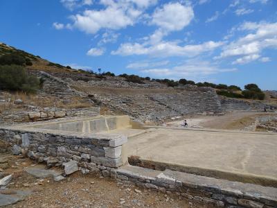 The image shows the ruins of an ancient stone theater with tiered seating carved into a hillside and a reconstructed concrete stage area in the foreground under a blue sky.
