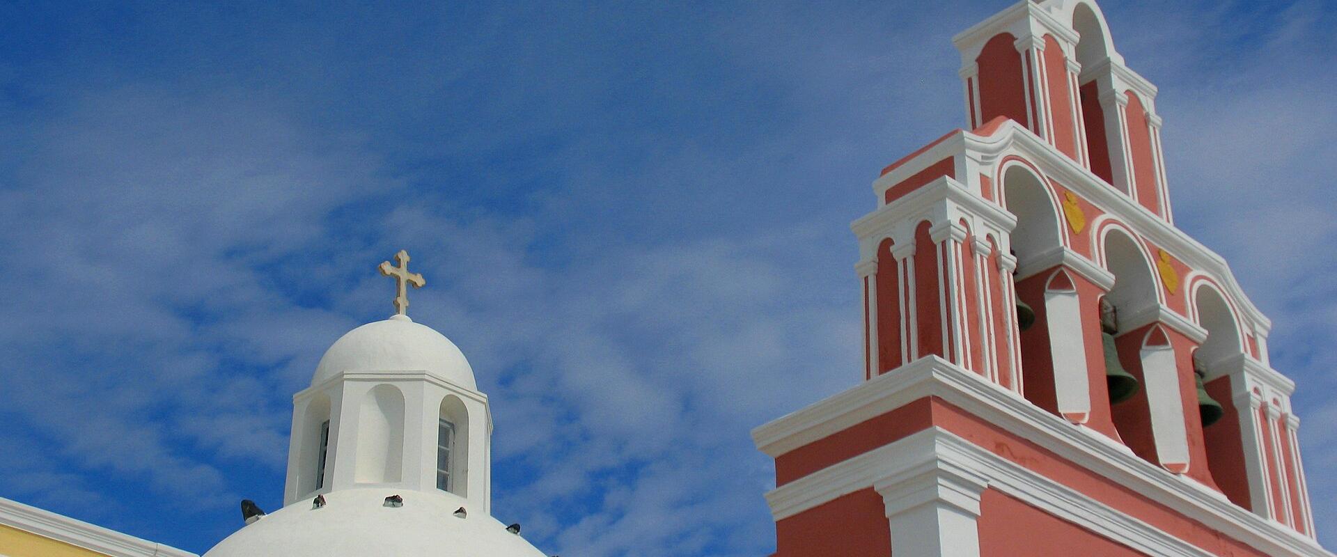 The image shows the upper portions of two church structures against a blue sky; the structure on the right is a tall, multi-tiered bell tower painted a reddish-pink color, while the structure on the left is a white dome with a cross.
