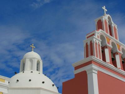 The image shows the upper portions of two church structures against a blue sky; the structure on the right is a tall, multi-tiered bell tower painted a reddish-pink color, while the structure on the left is a white dome with a cross.