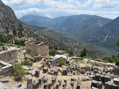The image shows ancient stone ruins on a mountainside with a view of a valley and distant mountains, featuring a prominent rectangular stone building (the Treasury of the Athenians) and scattered column bases.